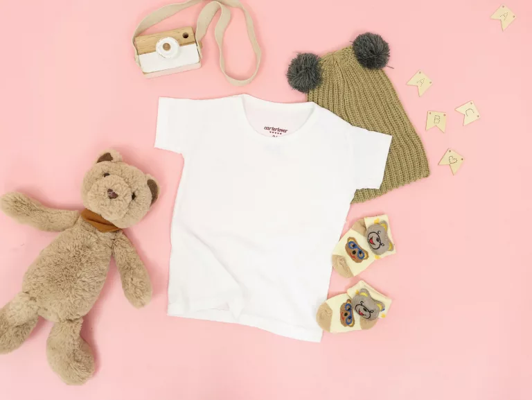Blank white baby clothes lying on a pink surface next to a teddy bear and a cute bear hat