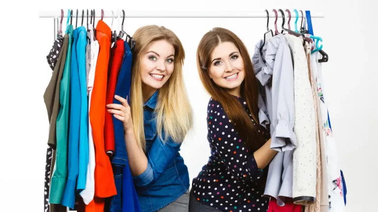 Off-Season Clothes Shopping - two women standing in front of a rack of clothing. They appear to be happy and possibly engaged in a fun shopping experience together. .