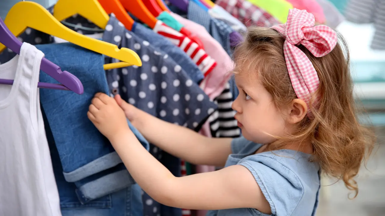 Save on Kids' Clothes - a girl, looking at clothes on hangers. The child has light brown hair styled with a pink and white gingham bow headband.