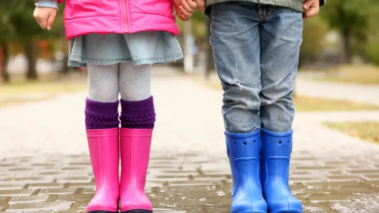 the lower bodies of two children holding hands, standing on a wet surface that suggests recent rain.