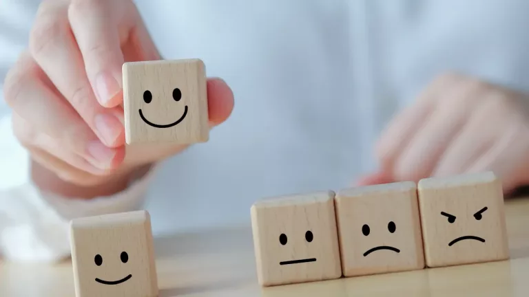 a person's hand holding a wooden cube with a smiling face drawn on it, positioned above three other wooden cubes placed on a surface in a row. Each of the cubes on the table also has a face.