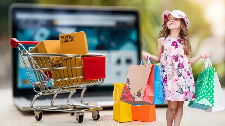 A mini shopping cart with boxes beside a laptop, and a young girl holding colorful shopping bags, symbolizing the contrast between online and in-person shopping.